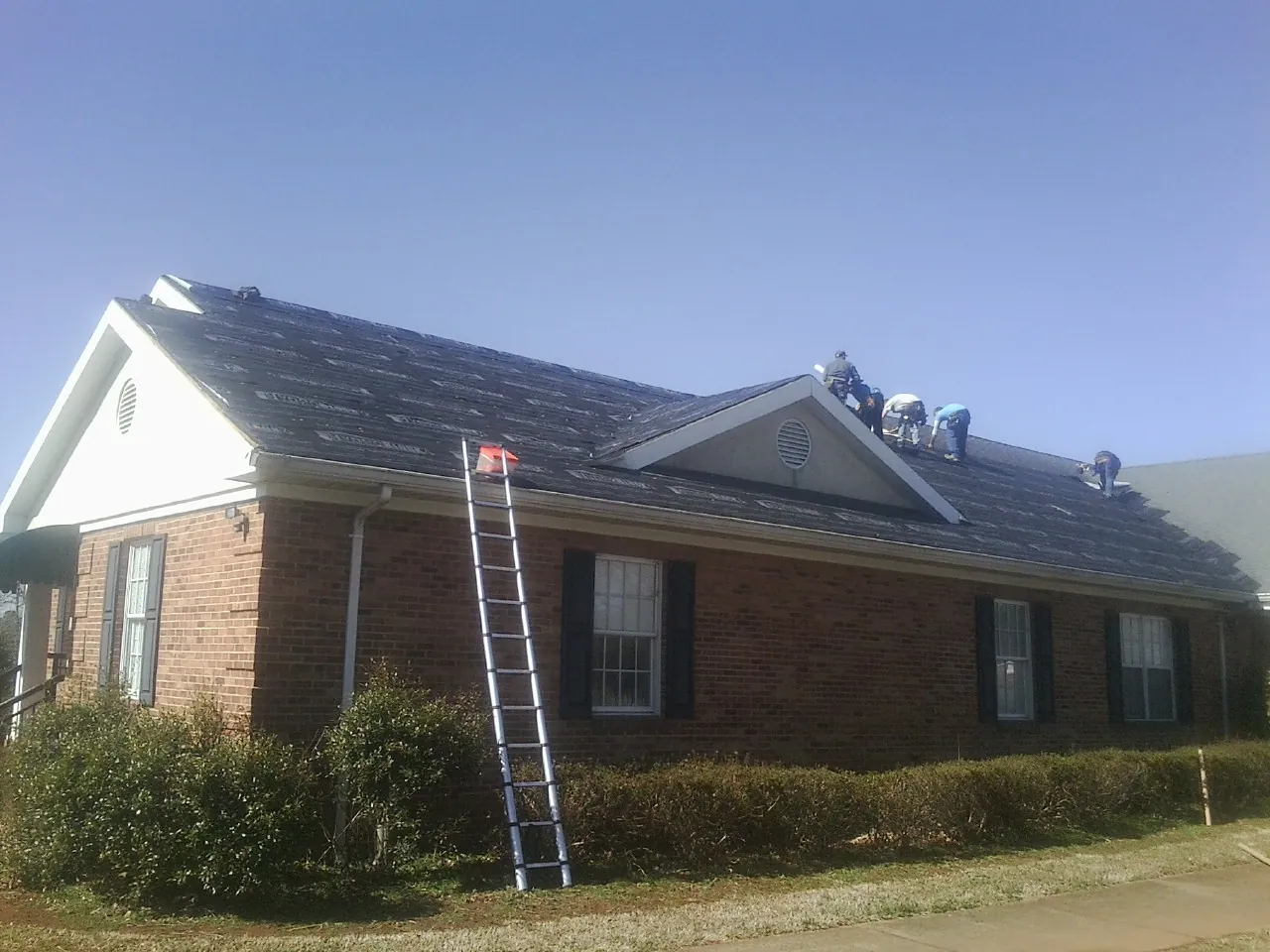 Crew tearing shingles off a brick ranch roof with a ladder set — Emergency Roof Repair work by our crew in Hitterdal